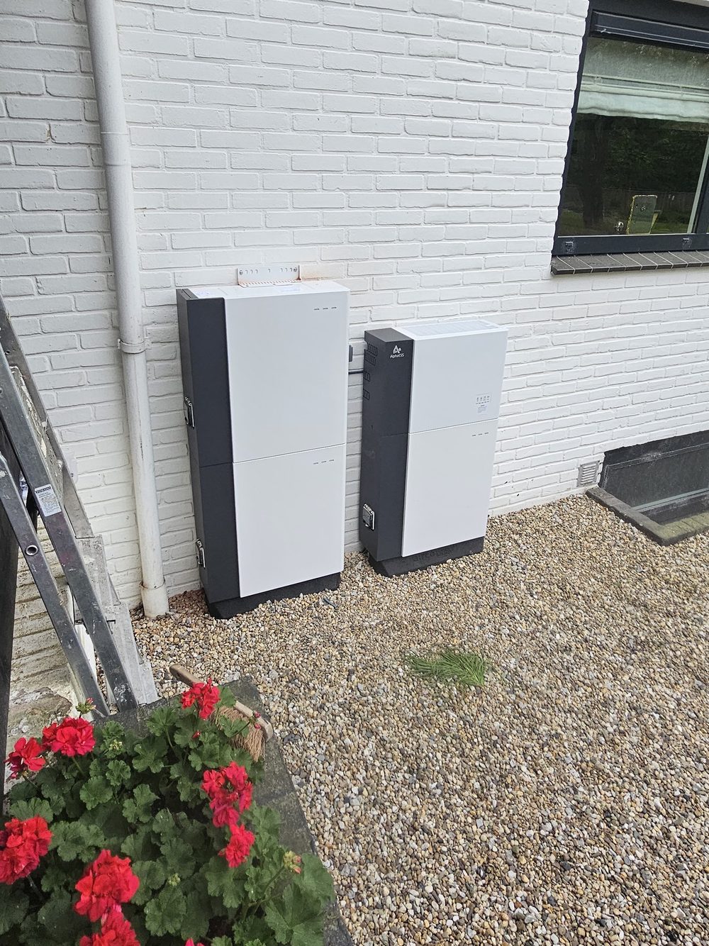 Twee thuisbatterijen tegen een witte gevel op grind, met rode geraniums